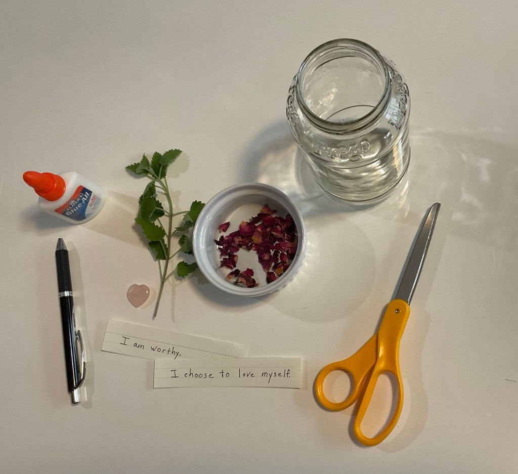 Items include jar, glue, catnip, rose petals, scissors, pen, heart shaped rose quartz crystal and affirmations on slips of paper saying “I am worthy” and “I choose to love myself.”