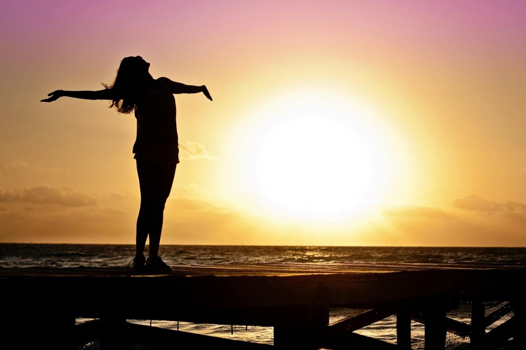 A woman at a dock on a body of water, arms stretched out and head back enjoying the sunrise.