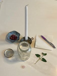 A jar with a life, a dish with rose petals, a white candle,a pen  and paper, a catnip sprig and a piece of rose quartz.